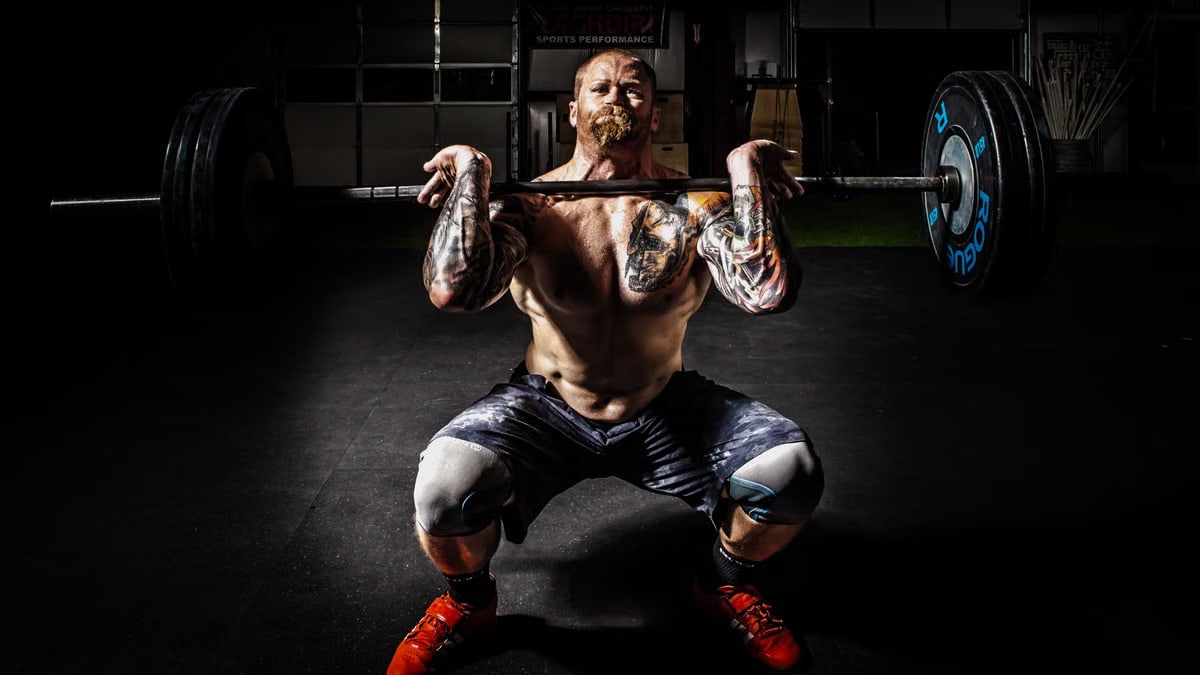 A tattooed man performing a heavy barbell squat in a gym for strength training.