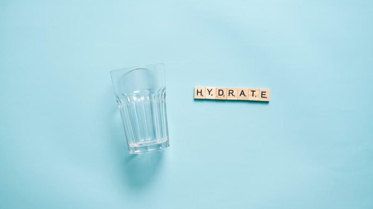 Minimalist flat lay of a drinking glass and letter tiles spelling 'HYDRATE' against a blue background.